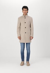 Abrigo de lana beige con cuello alto, cinco botones negros y bolsillos laterales, llevado sobre jeans azules y zapatos beige. Textura suave y diseño ajustado.