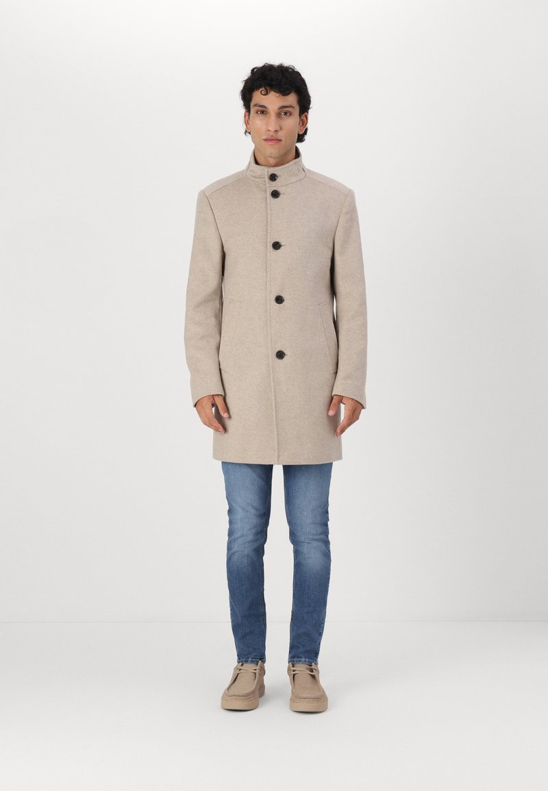 Abrigo de lana beige con cuello alto, cinco botones negros y bolsillos laterales, llevado sobre jeans azules y zapatos beige. Textura suave y diseño ajustado.