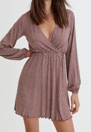 Femme portant une mini robe à manches longues, col en V profond, avec un motif ondulé à points marron et blancs, taille élastique et ourlet volanté, sur fond uni.