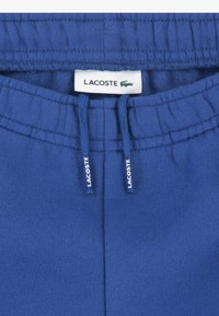 Sinised puuvillased dressipüksid, millel on elastne vöökoht paeltega ja tekstuuriga viimistlus. Lacoste'i logo on nähtav vöökohal.