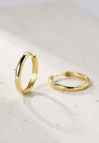 Goldene Creolen mit glatter, glänzender Oberfläche, die eine kreisförmige Form und ein leichtes Scharnier für den Verschluss aufweisen, vor einem hellen Hintergrund präsentiert.