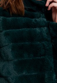 Měkká, tmavě petrolejová umělá kožešina s prošívaným vzorem, charakterizovaná plyšovou texturou a viditelnými švy zvýrazňujícími nadýmané části.