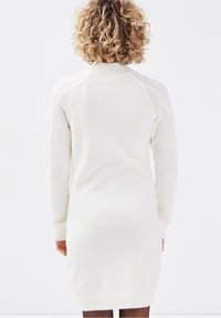 Personne aux cheveux frisés portant une robe en tricot blanche avec des motifs en câble et texturés sur les manches, se tenant de dos face à la caméra.