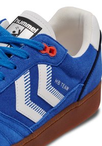 Baskets en suède bleu avec un design en chevron blanc, une boucle de lacet rouge et une semelle en caoutchouc naturel. Présente une étiquette logo noir et blanc sur la languette.