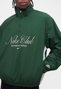Άνδρας που φοράει σκούρο πράσινο πουλόβερ Nike Club International με λευκό λογότυπο και φερμουάρ στον γιακά, τα χέρια στις πλαϊνές τσέπες.