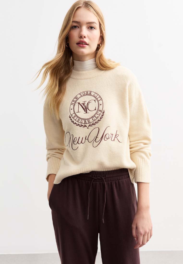 Beige Pullover mit hohen Kragen, versehen mit einem runden Logo und dem Text "New York" in kastanienbraun. Kombiniert mit dunkelbraunen, mit Kordelzug versehenen Hosen.