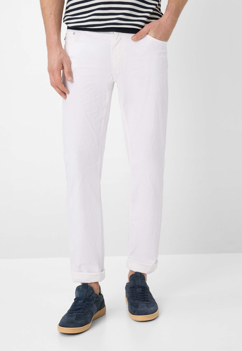 Homme portant un pantalon blanc retroussé, des baskets bleu marine à semelles en gomme et une chemise rayée noir et blanc, debout avec une main dans la poche.