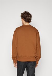 Sudadera marrón con cuello redondo, ribete en el dobladillo y hombros caídos, combinada con pantalones negros. La tela parece suave con una textura sólida.