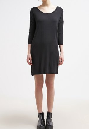 Femme portant une robe noire simple longueur genoux avec des manches trois-quarts et des bottines noires, debout sur un fond blanc uni.