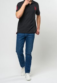 Černé polokošile s červeným logem na hrudi v kombinaci s modrými džínami a bílými teniskami. Model na neutrálním pozadí.
