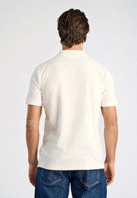 Vit bomullspikétröja med korta ärmar, en uppvikt krage och en texturerad yta. Bärs med blå denimjeans, vilket visar en avslappnad passform.