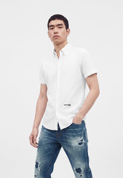 Jeune homme portant une chemise blanche à manches courtes boutonnée et un jean bleu déchiré, debout avec une main dans la poche.