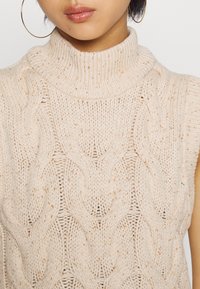 Maglione smanicato color beige con collo alto, caratterizzato da un motivo a treccia testurizzato e sottili puntini. Materiale leggero e morbido con un taglio morbido.
