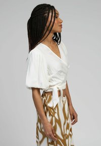 Haut blanc cache-cœur avec manches ballon, noué à la taille ; associé à une jupe marron ornée d'un motif abstrait blanc.