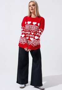 Czerwony sweter z dzianiny z białymi wzorami w serca, geometrycznymi wzorami i prążkowanym wykończeniem. Połączony z szerokimi czarnymi spodniami i trampkami.