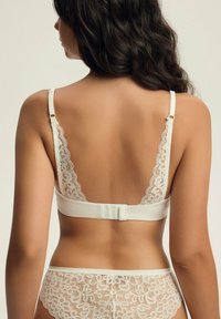 Biały koronkowy stanik z gładkim paskiem materiałowym, z wyszukanym koronkowym wykończeniem na ramiączkach oraz zapięciem na haftki z tyłu.