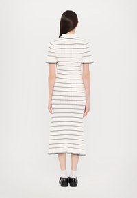 Robe rayée côtelée en crème avec des lignes marine, à manches courtes et avec un ourlet évasé. Associée à des chaussures noires et des chaussettes blanches. Vue de dos.