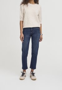 Jersey de punto beige con mangas texturizadas, combinado con jeans azul oscuro y zapatillas claras. Las zapatillas tienen cordones negros y detalles en color marrón claro.