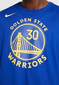 Modré bavlněné tričko s krátkými rukávy. Na přední straně má kruhovou grafiku: "Golden State Warriors," "30" a ilustraci mostu ve žluté barvě.