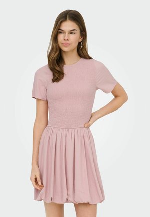 Jeune femme aux longs cheveux bruns portant une robe courte à manches courtes rose clair, smockée avec une jupe froncée, debout avec une main sur la hanche.