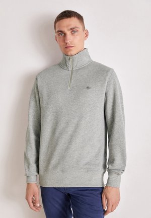 Jeune homme aux cheveux courts portant un sweat-shirt gris clair avec une fermeture éclair jusqu’au quart et un pantalon bleu foncé, debout devant un fond clair uni.