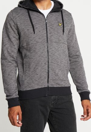 Mann trägt grauen Kapuzenpullover mit Reißverschluss, schwarzer Kapuzenfütterung, schwarzen Kordeln, gerippten Bündchen und Saum sowie einem kleinen gelben gestickten Logo auf der Brust.