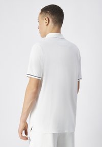 Camisa polo de algodón blanco con un diseño liso, presenta acentos de raya negra en las mangas y aberturas laterales cerca del dobladillo.