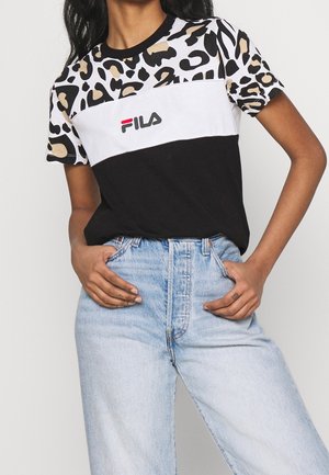 Osoba ubrana w czarno-biało-panterkowy t-shirt FILA oraz jasnoniebieskie, wysokowalne jeansy, z rękami w przednich kieszeniach.