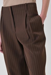 Pantaloni marroni a righe sottili realizzati in tessuto liscio, con pieghe, tasche laterali e vestibilità su misura. Una sottile lucentezza enfatizza la trama.
