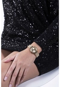 Kerek aranytone óra, textúrált arany szíjjal, amelyet tiszta rónai kövek díszítenek, fekete flitteres ruhával kombinálva.