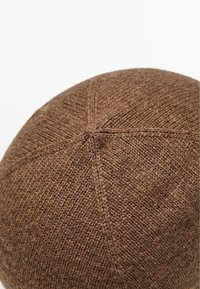 Bruine gebreide muts met een ronde top, voorzien van een textuurpatroon en cirkelvormige stikdetails op de kroon. Gemaakt van een zachte wolmix.