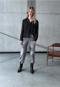 Camicia nera a bottoni abbinata a pantaloni grigi chiari e stivali alla caviglia neri. Sullo sfondo si vede un muro di cemento e una sedia di legno.