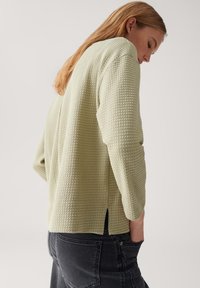 Donna con capelli lunghi che indossa un top a maniche lunghe di un verde chiaro a trama e jeans neri, con le mani in tasca, di profilo su uno sfondo neutro.