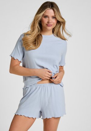 Giovane donna con lunghi capelli biondi che indossa un top azzurro chiaro a costine con maniche corte e pantaloncini abbinati, tenendo delicatamente l'orlo della maglietta.