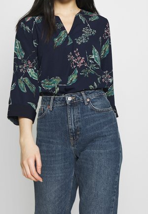 Vrouw die een marineblauw bloesje met bloemenprint en driekwartmouwen draagt, ingestopt in een jeans met hoge taille.