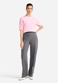 Maglione rosa a maniche corte in morbido tessuto a maglia, abbinato a pantaloni grigi su misura con cuciture verticali. Le calzature sono bianche con punta affusolata.