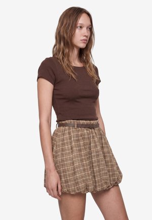 Femme aux cheveux châtain clair ondulés portant un haut marron ajusté à manches courtes et une jupe beige à carreaux taille haute avec ceinture, debout de profil.