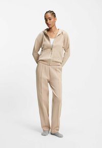 Sudadera beige con cremallera y bolsillo frontal, combinada con pantalones ajustados a juego. Ambas prendas presentan una textura suave y un ajuste informal.