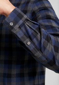 Primer plano de un brazo doblado que lleva una camisa de manga larga a cuadros en azul y gris con los puños abotonados.