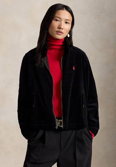 Γυναίκα με μακριά σκούρα μαλλιά που φοράει λευκά σκουλαρίκια, κόκκινο ζιβάγκο, μαύρο μπουφάν με φερμουάρ και κόκκινο λογότυπο, και μαύρο παντελόνι, στέκεται μπροστά σε ουδέτερο φόντο.