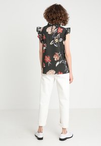 Blusa a fantasia floreale in tessuto scuro con maniche arricciate, caratterizzata da vivaci disegni di fiori. Abbinata a pantaloni bianchi arrotolati e scarpe nere.