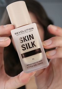 Revolution Skin Silk Serum Foundation in einer frosted klaren Flasche mit einem beigen Deckel. Der Text zeigt den Produktnamen, die Nuance "F2" und Details in Schwarz.