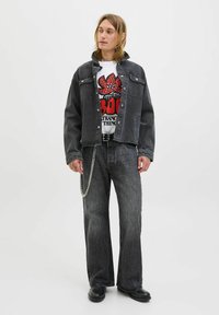 Jachetă din denim negru purtată peste un tricou alb cu grafică, asortată cu blugi negri cu picior larg. Caracteristicile includ un accesoriu cu lanț și cizme negre.