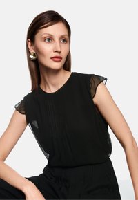 Schwarze Bluse mit kurzen, transparenten Ärmeln, mit plissierten Details vorne. Einfacher Ausschnitt, weicher Stoff, tailliert geschnitten. Silberne Ohrringe.