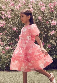 Roza obleka s puhastimi rokavi, cvetličnim vzorcem, večstopenjskim krilom in vodoravnimi gubami. Nošena z belimi sandali. V ozadju zelena listnata rastlina in rožnate cvetlice.