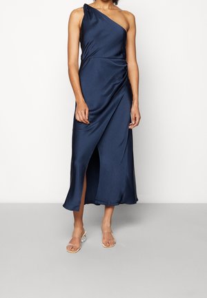 Granatowa sukienka maxi z satyny z jednym odsłoniętym ramieniem, bocznym rozcięciem i marszczonym górnym dołem. Do tego przezroczyste sandały na obcasie.