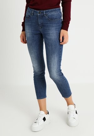 Woman wearing cropped blue skinny jeans, white platform sneakers, and a burgundy long-sleeve top, standing against a plain white background.