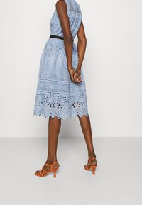 Kvinna som bär en knälång ljusblå spetsklänning med geometriska mönster och orange högklackade sandaler, stående med händerna bakom ryggen.