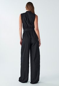 Črn skok z odprtimi rokavi z ovratnikom, poudarjenim pasom in širokimi hlačnicami. Gladek, lahek material z rahlo teksturo.