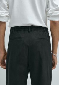 Zwarte broeken met elastische tailleband, twee achterzakken, zachte textuur en rechte snit. Draagt een witte blouse met lange mouwen.